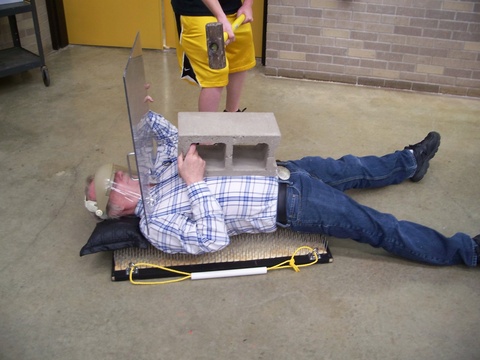 person wearing face protection, plexiglass plate laying on a bed of nails with block on chest, person holding a sledgehammer